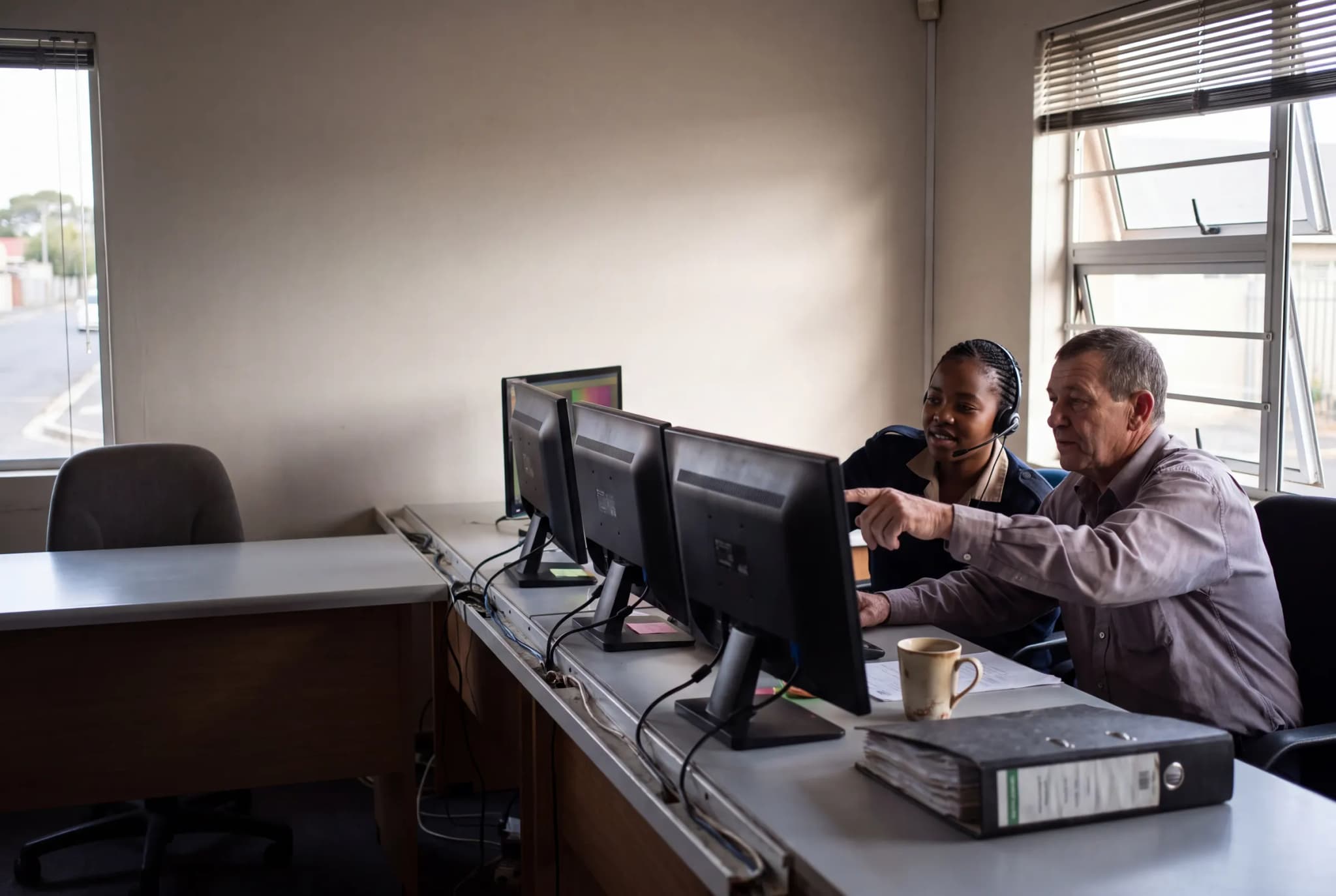 A control room operator wearing a headset and a supervisor looking at the same monitor together